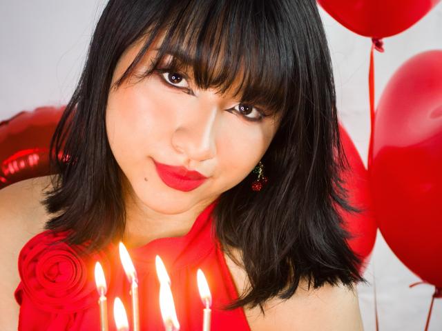 DariaHotst with black hair, red lipstick, and red dress, holding candles in front of her, surrounded by red balloons. Her tits are slightly visible under the dress.