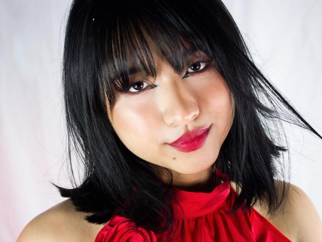 DariaHotst has black hair, red lipstick, and a small mole on her right cheek, wearing a red halter top. She's looking at the camera with a slight smile.