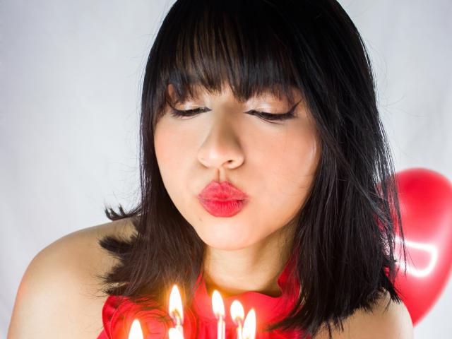 DariaHotst with black bob, red lipstick, closed eyes, pouty lips, holding candles, red scarf, red balloon in background, soft light, slight blush on cheeks.