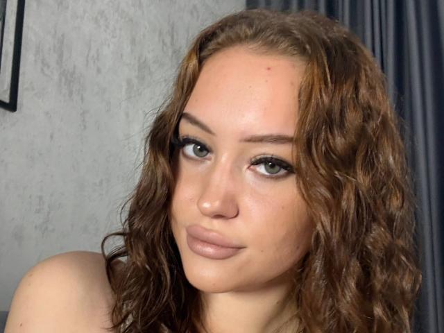 Michele Jinkins tem cabelos castanhos ondulados, olhos verdes e lábios cheios e rosados, com um leve sorriso. Ela está olhando para a câmera com uma expressão confiante e sensual..