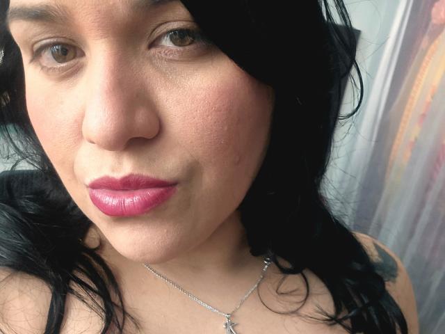VivianaThompson's face with pink lipstick, brown eyes, black wavy hair, and a silver necklace with a cross. She has a slightly flushed, textured skin and a subtle smile.