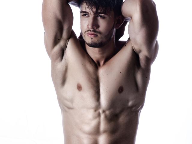 JordanPhillip, sin camisa, musculoso, con los brazos levantados, abdominales definidos, pezones pequeños, cabello corto y oscuro, barba incipiente, expresión seria, fondo blanco, cuerpo tonificado, piel clara..