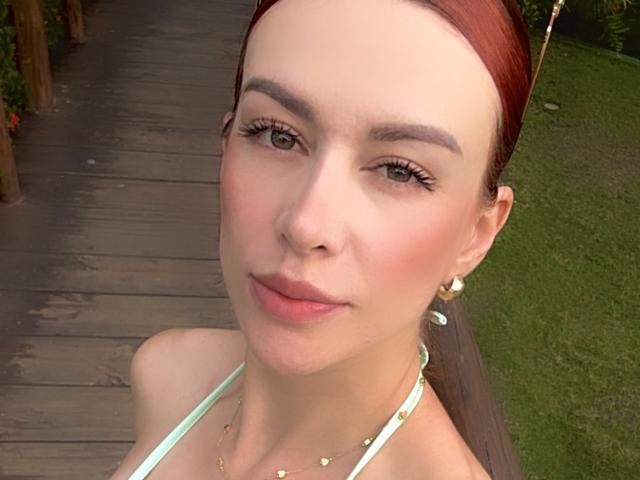 Leahred has pale skin, red hair in a ponytail, green eyes, and is wearing a green halter top with gold jewelry. She has a subtle smile and light makeup.