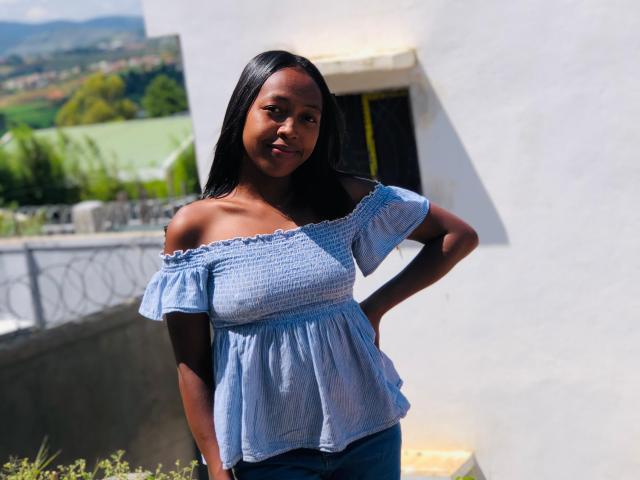 QueenBella69 with dark skin, long black hair, small tits, wearing a blue off-shoulder top and blue jeans, standing outside with hands on hips, smiling.