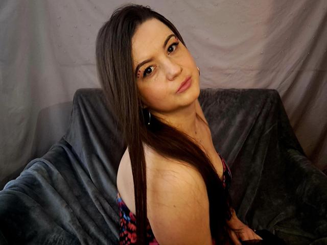 MavkaCute with long brown hair, nose ring, and subtle makeup, looking over her shoulder, wearing a colorful patterned top, sitting on a dark, textured chair.