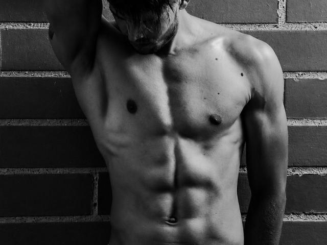 Il torso muscoloso e senza camicia di JordanPhillip, con addominali definiti, piccoli capezzoli e una leggera ombra sul lato destro. Bianco e nero, muro di mattoni sullo sfondo. In posa con un braccio alzato..