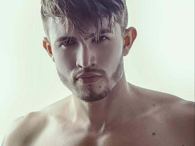 JordanPhillip ha un'aria seria, capelli castani, pelle chiara e una barba con un leggero baffo. Le sue spalle scoperte mostrano alcuni piccoli nei. È senza camicia e guarda in basso..