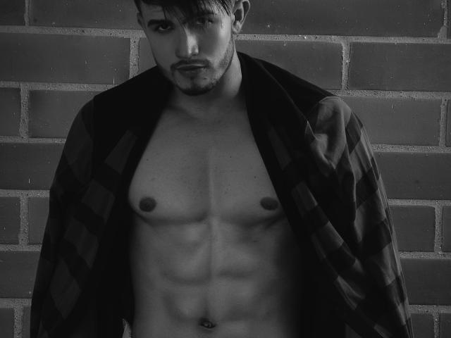 JordanPhillip, shirtless, with a plaid jacket open, exposing his toned abs and small, dark nipples. He has short, dark hair, a serious look, and a slight beard. Black-and-white.