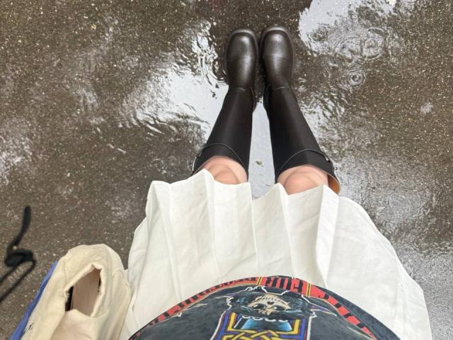 AnneKofron's legs in black knee-high socks and brown boots, standing in a puddle. She's wearing a white pleated skirt and a colorful top with a patterned design.
