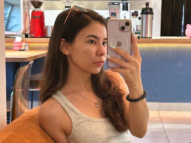 AnneKofron with long brown hair, wearing a white tank top, taking a mirror selfie in a cafe, showing her small tits, holding a phone with a white case.