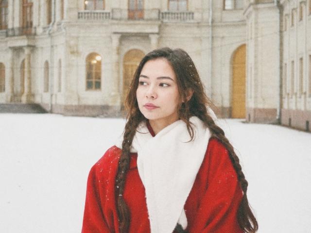 AnneKofron has long brown hair, a red coat, and a white scarf, standing in snow with a serious expression, snowflakes falling on her face.