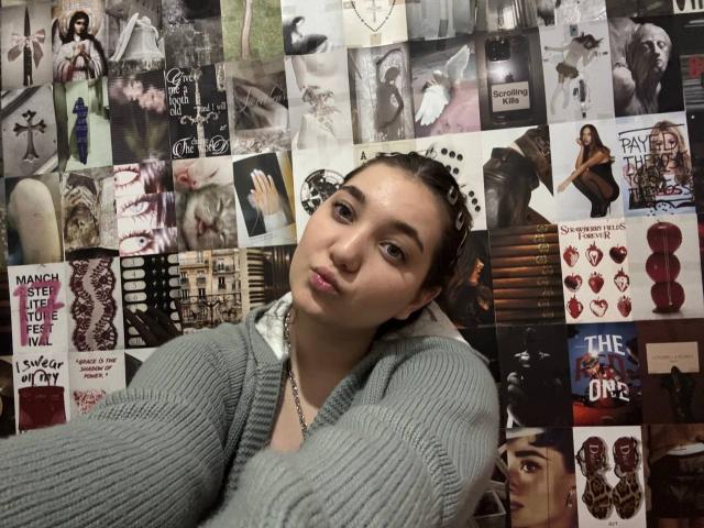 MichikoVivona with pouty lips, wearing a gray ribbed sweater, standing against a wall of black-and-white and color photos, has brown hair in small curls, light skin.