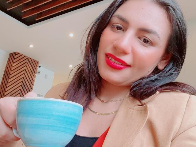 CamilleRiver with pink lipstick, holding a blue ceramic cup, wearing a tan jacket, black top, and gold necklace, smiling at the camera.