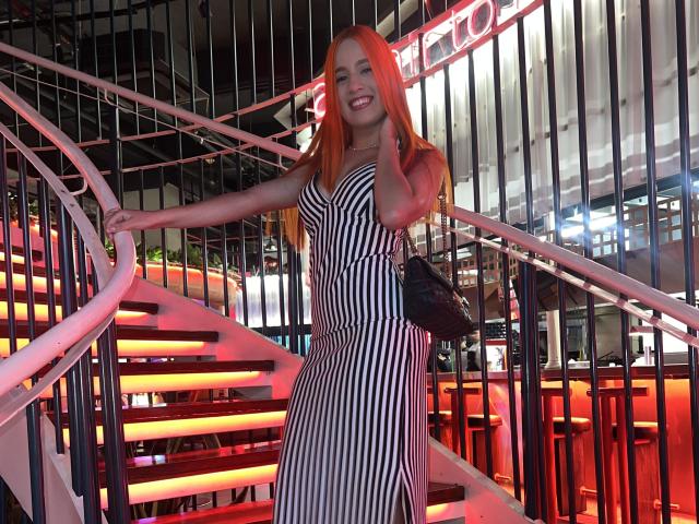 EmmaWat with long orange hair, black-and-white striped dress, holding black purse, standing on curved staircase, smiling, hands on railing, neon lights in background, nighttime setting.