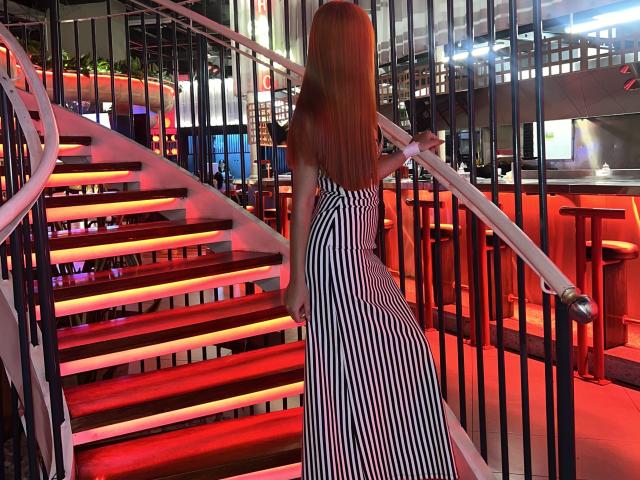 EmmaWat with red hair, striped dress, climbing red-lit stairs, holding rail, facing away, showing her ass. Indoor bar setting, red and black colors, modern decor.