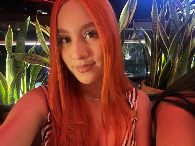 EmmaWat with bright red hair, cute smile, and pink glossy lips, wearing a striped top, sitting near green potted plants, in a dimly lit, cozy bar.