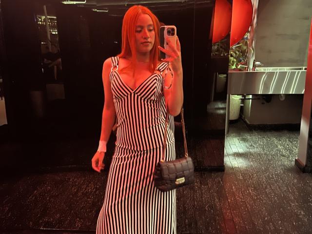 EmmaWat with red hair, wearing a black and white striped dress, holding a gray quilted purse, taking a mirror selfie with her phone, showing her tits and ass.
