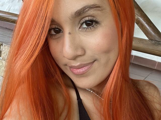 EmmaWat with orange hair, brown eyes, and pink lips, smiling at the camera, showing her cute nose and black halter top, with a small beauty mark on her left cheek.
