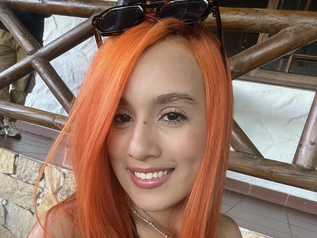 EmmaWat has bright orange hair, brown eyes, and a cute smile with white teeth. She's wearing sunglasses on her head, and has a silver necklace. Her ass is not visible.