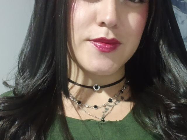 MiasXLovelys Gesicht mit glänzenden, rosafarbenen Lippen, schwarzem, glattem Haar, einem grünen Oberteil, einer schwarzen Kette mit Herzanhänger und zwei silbernen Ketten mit schwarzen Perlen. Zarte, helle Haut..
