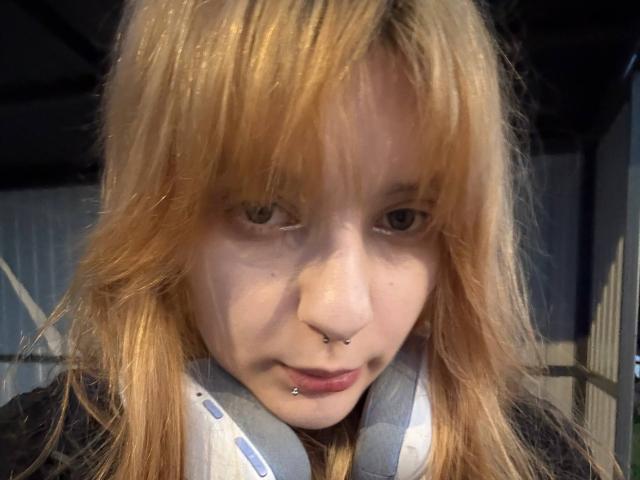 ForenCostler has blonde hair, a nose ring, and a lip piercing. She's wearing headphones with a blue and white strap, and looking down with a slight frown.