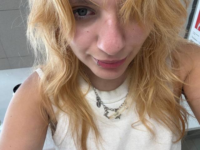 ForenCostler hat zerzaustes blondes Haar, blaue Augen und einen pinken Lippenring. Sie trägt ein weißes Rippstrick-Tanktop mit einer Halskette. Ihre Brüste sind nicht sichtbar..