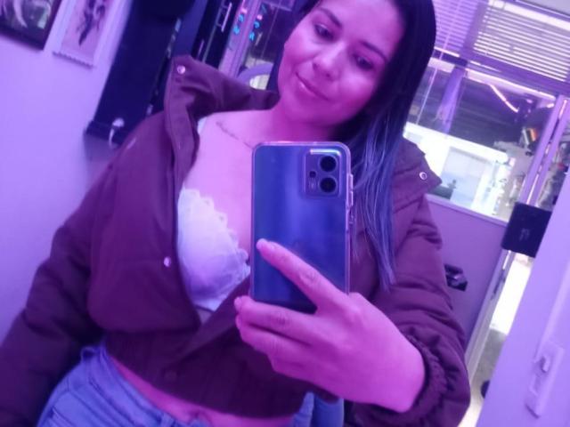 Nahoset with light brown skin and long dark hair, wearing an open brown jacket showing white lace bra, taking a selfie in a purple-lit room, holding a blue phone.