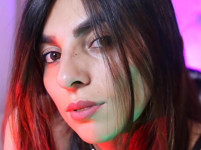 Le visage de SoyVeronica, avec ses cheveux bruns ondulés, ses lèvres légèrement entrouvertes et un œil fermé, est illuminé par des lumières rouges et vertes. Son nez est petit, et elle a une peau claire..