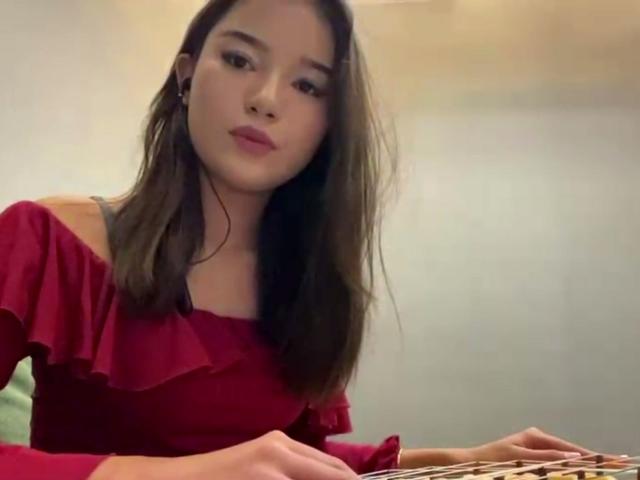 ZulaGullick, con lunghi capelli castani, una maglia rossa a spalle scoperte e rossetto rosa, seduta a un tavolo, che guarda leggermente di lato, con un'espressione calma e uno sguardo malinconico..