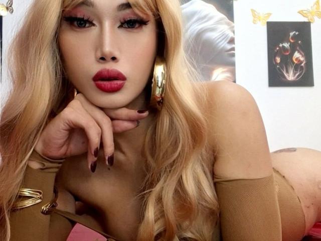xGoddessJazzyx, com cabelos loiros longos, batom vermelho, brincos de ouro grandes e mangas bege, deitada, com a mão no rosto, mostrando os seios e uma tatuagem no bumbum..