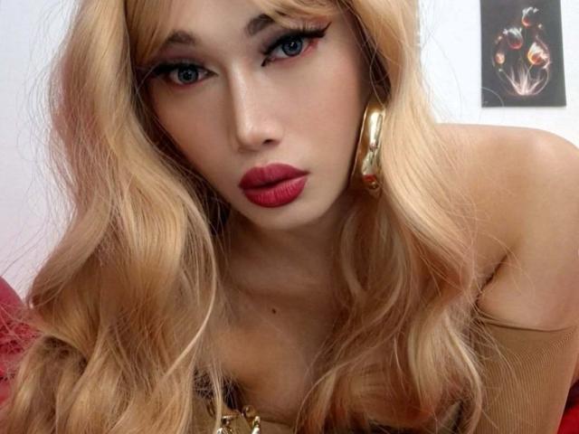 xGoddessJazzyx mit blonden Haaren, blauen Augen, rotem Lippenstift, goldenen Ohrringen und einem braunen Oberteil, die verführerisch in die Kamera schaut, wobei ihre Brüste und ein süßes, sexy Gesicht zu sehen sind..