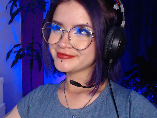 RavenThuwu with purple hair, glasses, red lipstick, nose piercing, chin piercing, headset, and blue shirt, smiling at camera. Boobless, no visible ass or pussy.