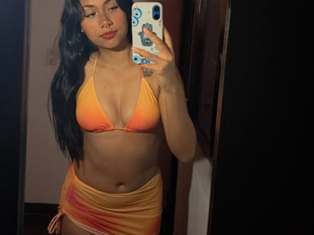 AnaRome with long black hair, orange bikini top showing her tits, and yellow-orange skirt, taking a mirror selfie, holding a phone with blue stickers, showing a small tattoo.
