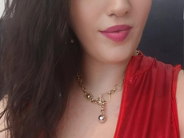 VanessaSquirtt with medium brown wavy hair, pink lipstick, red dress, and gold chain necklace with a dangling pearl. Her tits are partially visible.