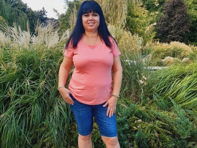 MatureVivian with black hair, pink shirt, and blue shorts, standing with hands on hips, showing her ass in a green garden. She's smiling, wearing a bracelet.