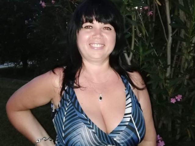 MatureVivian has black hair, brown eyes, and a smile. She's wearing a blue patterned dress with a deep V, showing her large boobs. She's standing outside at night.