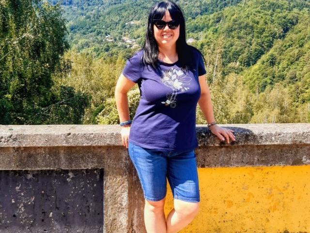 MatureVivian with black hair, sunglasses, blue shirt, and blue shorts, standing against a yellow and gray concrete wall, smiling. Her tits are medium-sized, and she's looking cute.