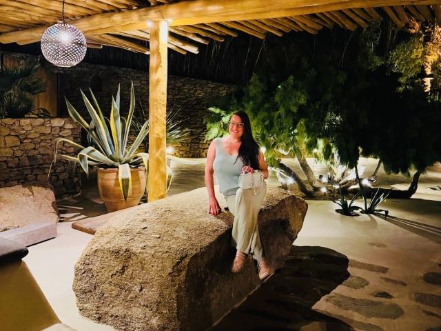 Ein BadAngel mit langen braunen Haaren, einem weißen Tanktop und beigen Hosen, sitzt auf einem großen Felsen, unter einer hölzernen Pergola, und hält eine weiße Handtasche, wobei eine Hängelampe zu sehen ist..