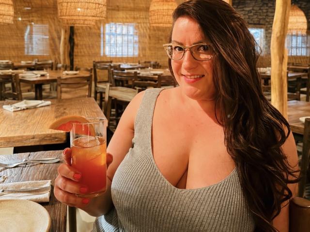 Uma mulher chamada BadAngel, com seios grandes, usando óculos e uma camiseta cinza de malha, segurando uma bebida, sorrindo em um restaurante de madeira, tem cabelos castanhos longos e uma figura curvilínea..