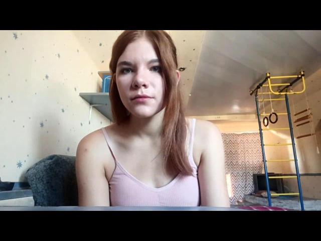 NeldaBase with red hair, pale skin, and small tits wearing a pink ribbed tank top, sitting at a table, looking serious, in a room with a yellow ladder.