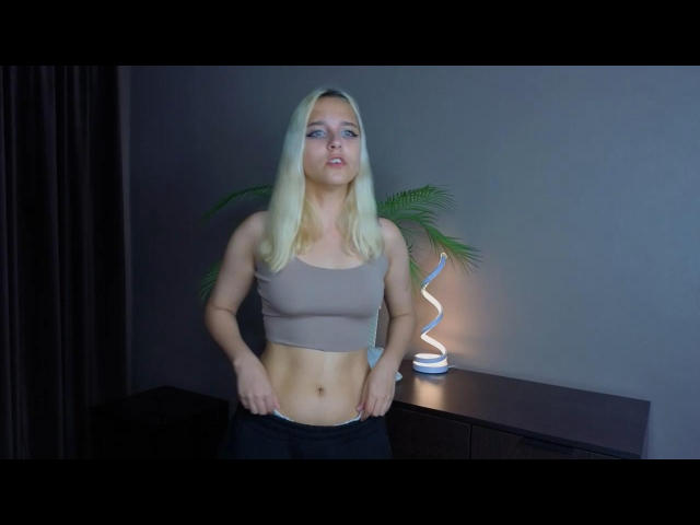 LouannMathai with blonde hair, blue eyes, and a tan sports bra, pulling down black pants to reveal her white underwear and toned abs, standing next to a lit lamp.