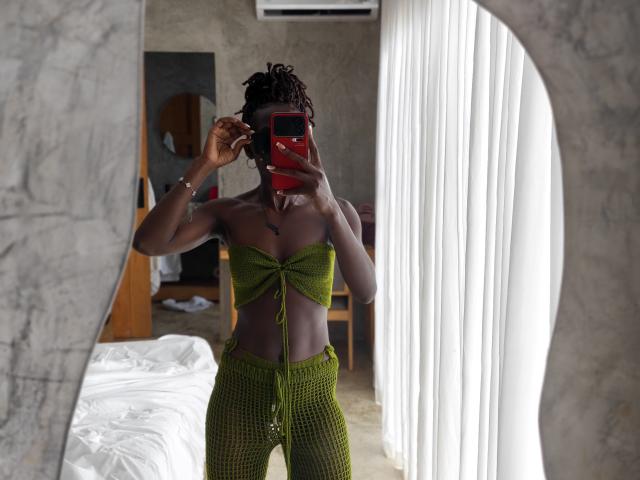 Glorydays with dark skin, small green knit top, tied just above the tits, and matching green knit pants, taking a selfie in a mirror, holding a red phone.