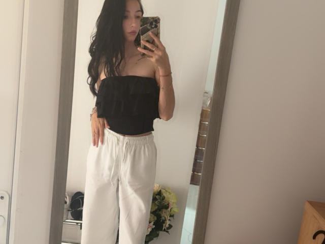 SinReea with long black hair, off-shoulder black top, and white pants, taking a mirror selfie, showing her boobs and ass in the reflection. Her phone has a floral case.