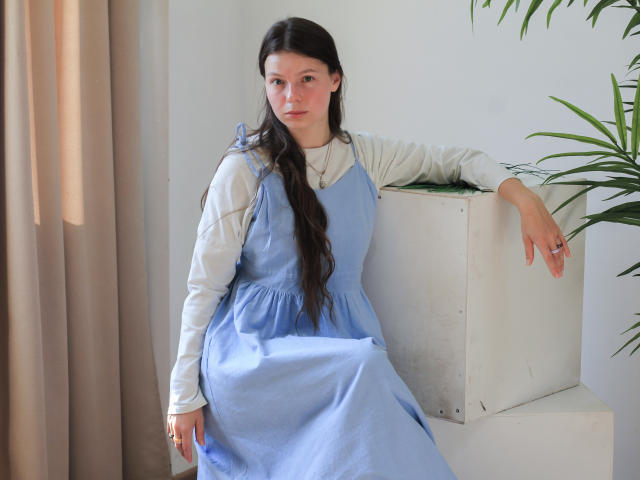 Jasmin, con lunghi capelli scuri, indossa un abito azzurro chiaro sopra una camicia bianca, appoggiata a una scatola bianca, con il braccio destro appoggiato, la mano sinistra sul ginocchio, e guarda verso la fotocamera..