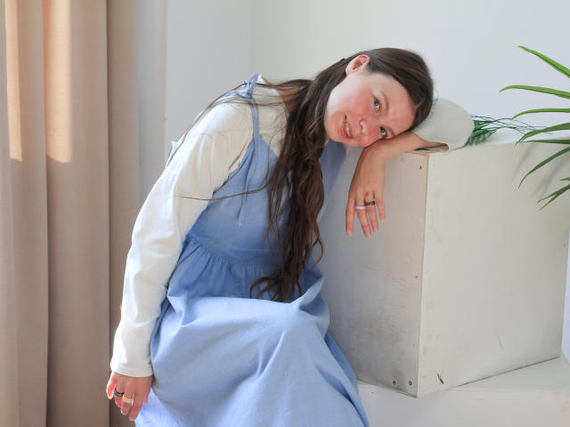 JasminHoy mit langen, braunen Haaren, einem weißen, langärmeligen Hemd und einem blauen Kleid, die sich an einen weißen Block lehnt, lächelnd, mit einem Ring am Finger, neben einer grünen Pflanze..