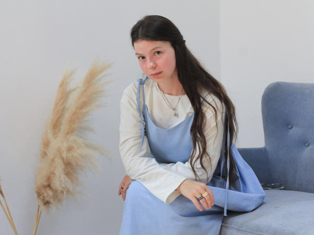 Jasmin, heute mit langem, dunklem Haar, in einem blauen Kleid über einem weißen Hemd, sitzt auf einem blauen Stuhl, hält trockenes, hellbraunes Gras in der Hand, trägt einen Ring und eine Halskette, blickt leicht nach unten und lächelt sanft..