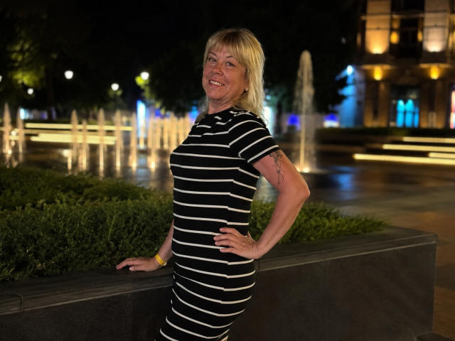ChristinaDavedes stands with hands on hip, wearing a black and white striped dress, smiling, blonde hair, small tits, tattoo on right arm, night, fountain in background.
