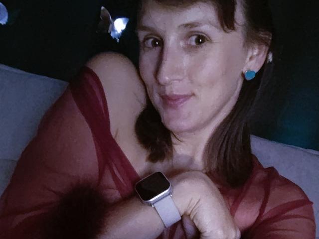FavoriteHare with medium tits, wearing a sheer red top, blue earrings, and a white smartwatch, smiling at the camera, brown hair, dim lighting, casual selfie.