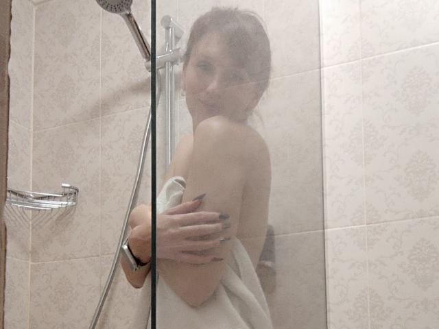 FavoriteHare in a glass shower, nude, with a towel covering her ass and tits, holding a showerhead, water droplets on the glass, light brown hair, smiling.