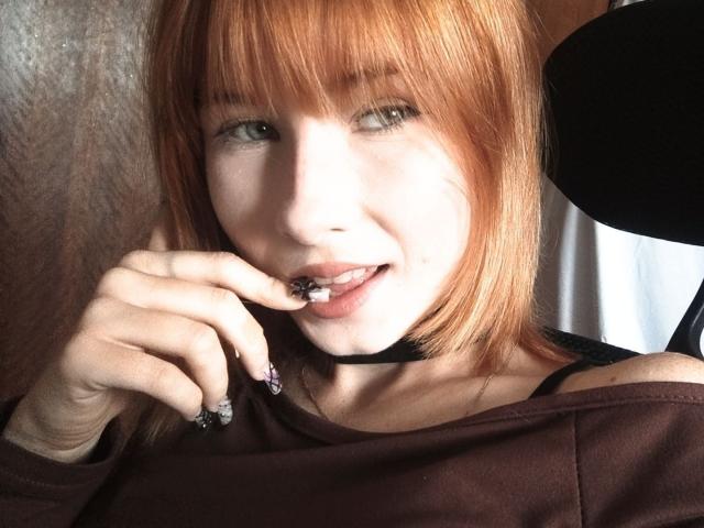 Kasandra Grothe mit roten Haaren, heller Haut und grünen Augen, die an einem schwarzen Lutscher lutscht, trägt ein braunes Hemd, eine schwarze Halskette und violette Schmetterlingsmotive auf den Nägeln..